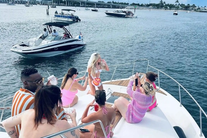 Group of people in swimwear relaxing on a boat with other boats in the background on a sunny day.