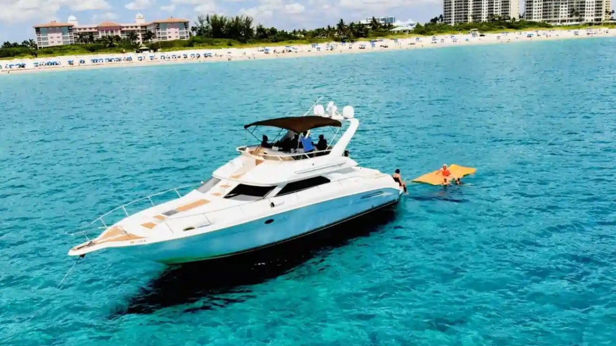 White yacht on blue water with people on a floating mat, beach and buildings in background.