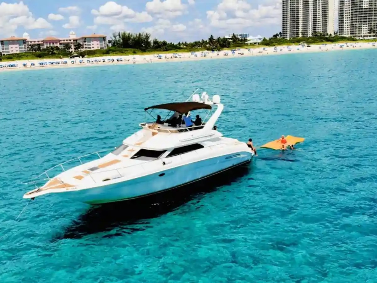 White yacht on blue water with people on a floating mat, beach and buildings in background.