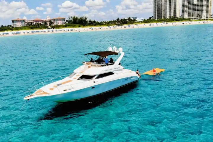 White yacht on blue water with people on a floating mat, beach and buildings in background.