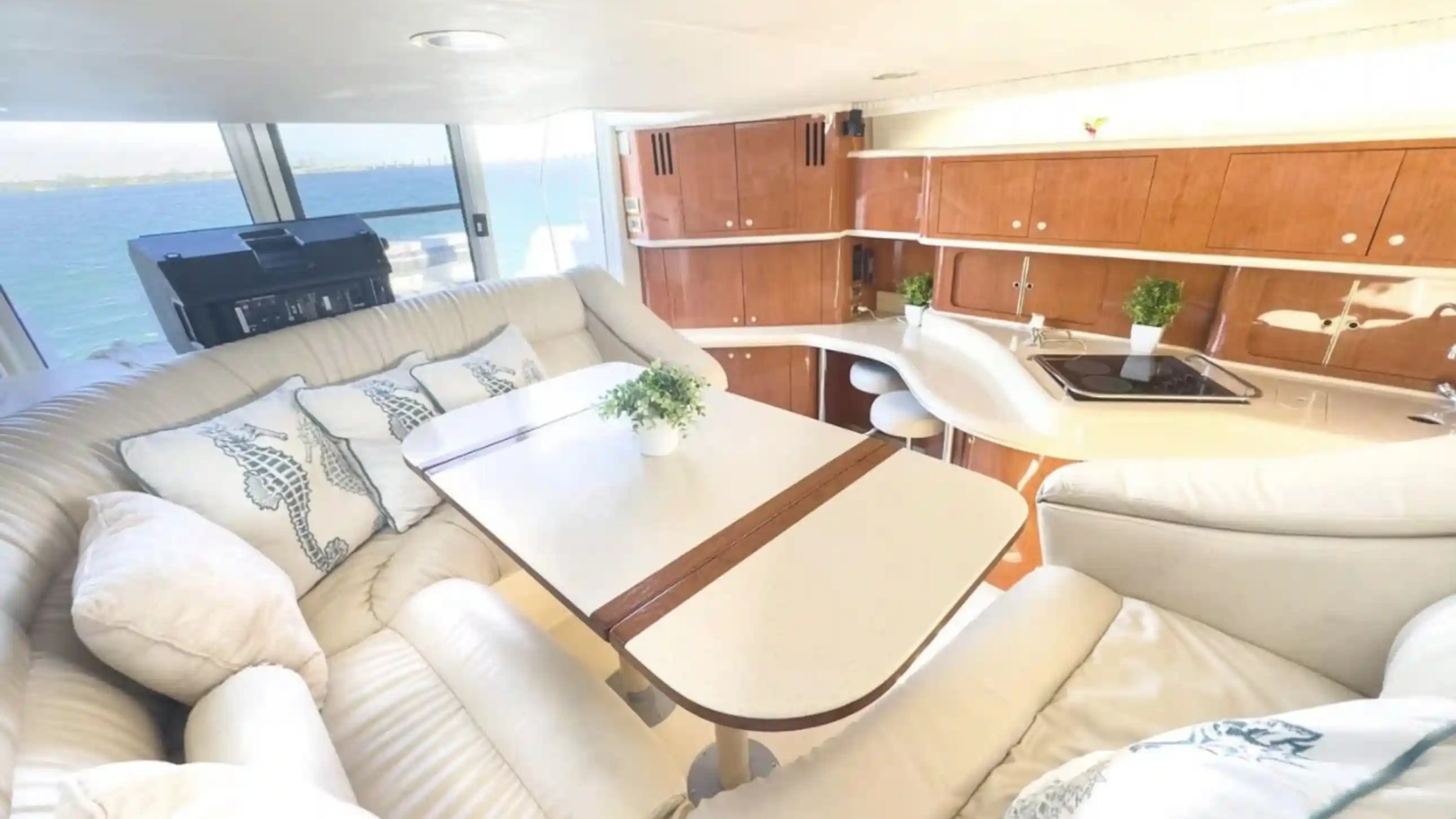 Interior of a yacht with beige seating, table, and kitchen area, overlooking the sea through large windows.