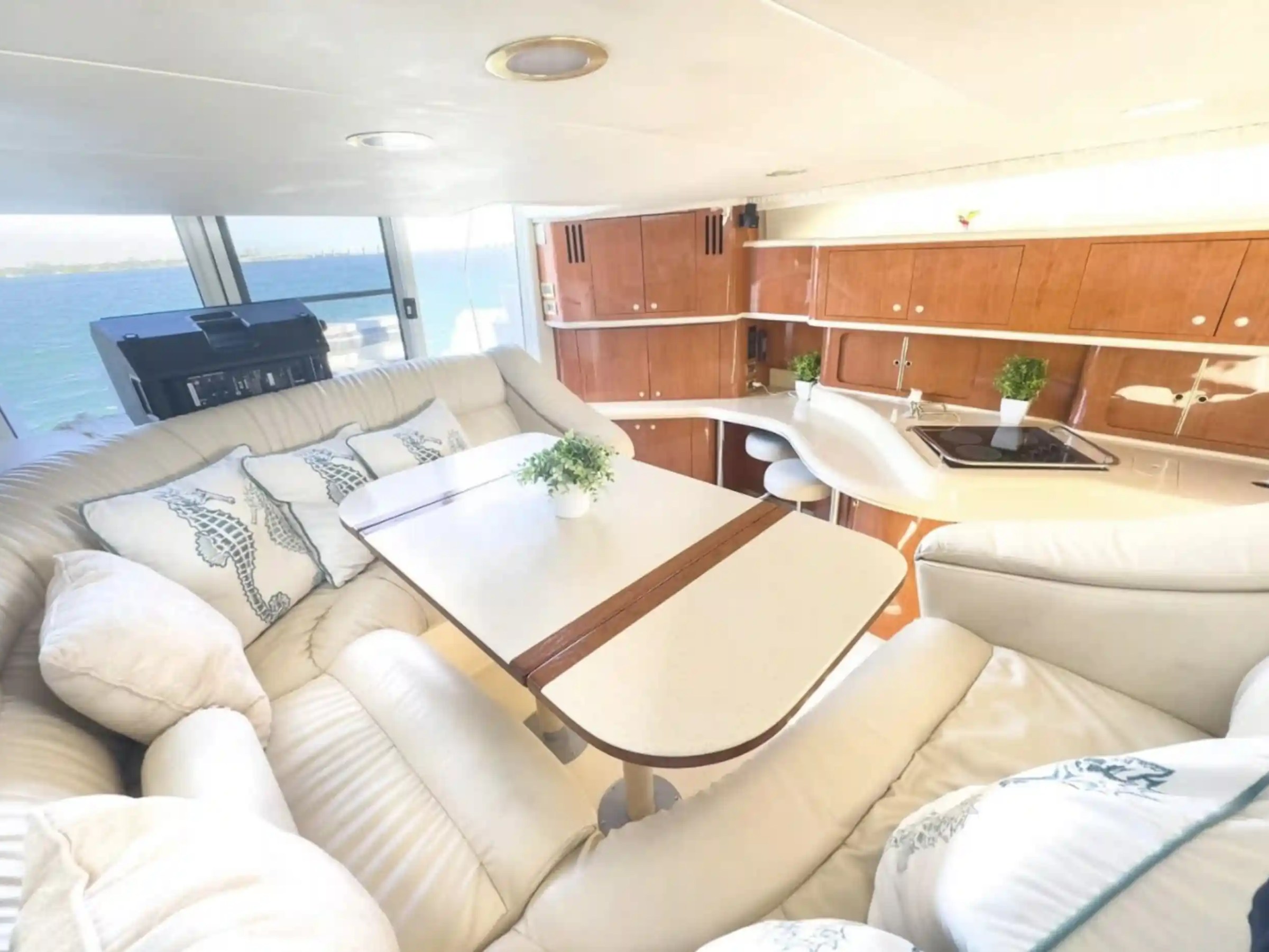 Interior of a yacht with beige seating, table, and kitchen area, overlooking the sea through large windows.