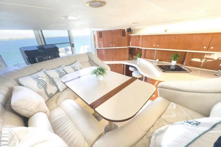 Interior of a yacht with beige seating, table, and kitchen area, overlooking the sea through large windows.