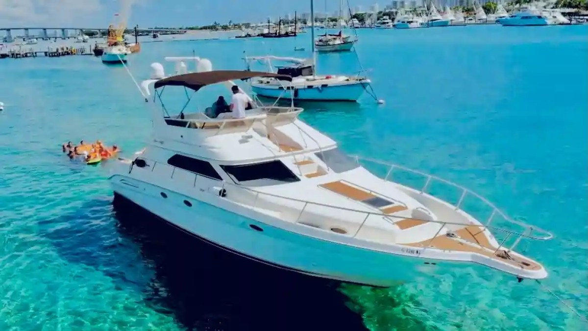 Yacht in clear blue water with people swimming nearby and boats in the background.