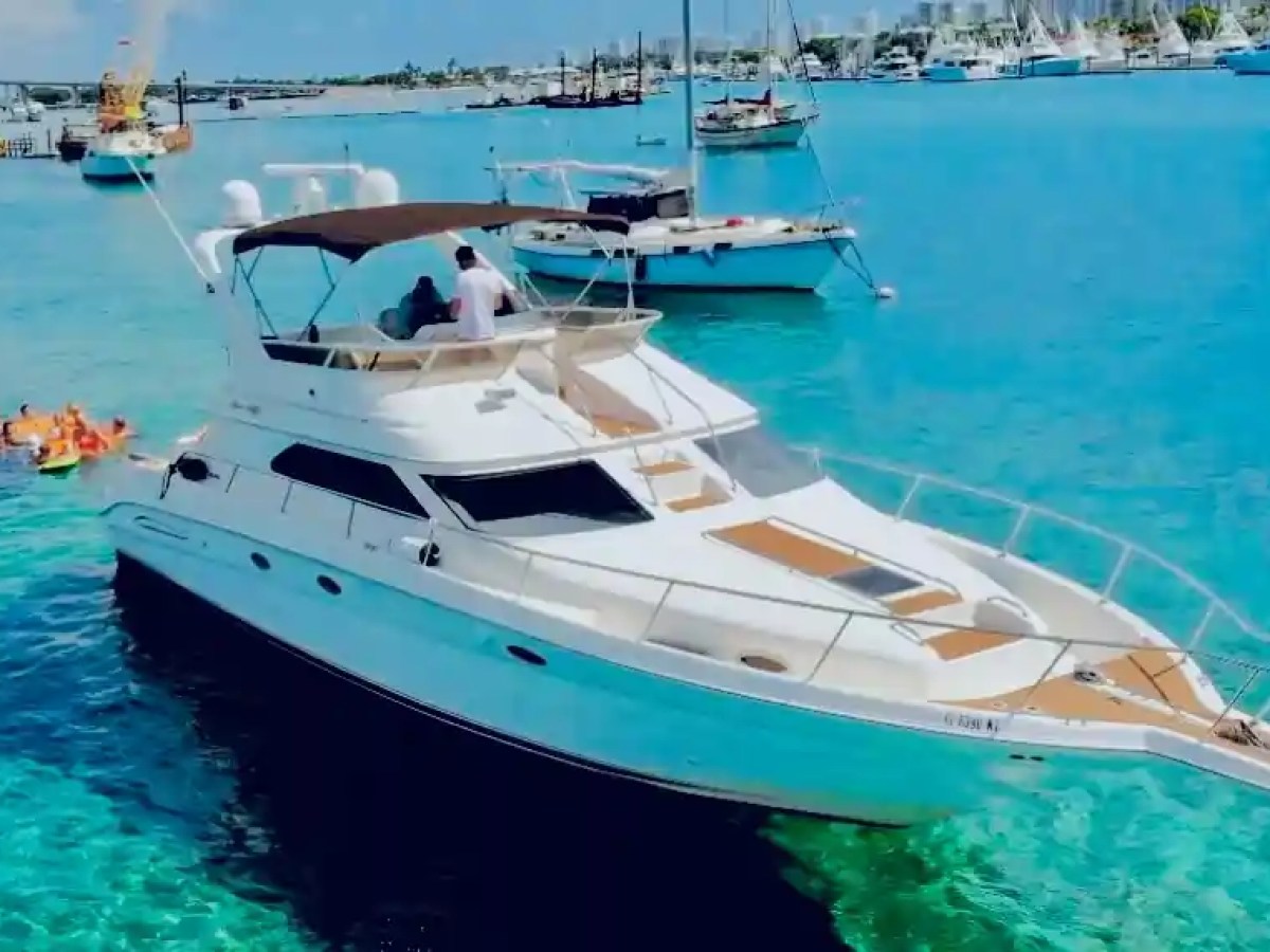 Yacht in clear blue water with people swimming nearby and boats in the background.