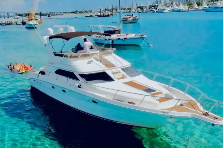 Yacht in clear blue water with people swimming nearby and boats in the background.