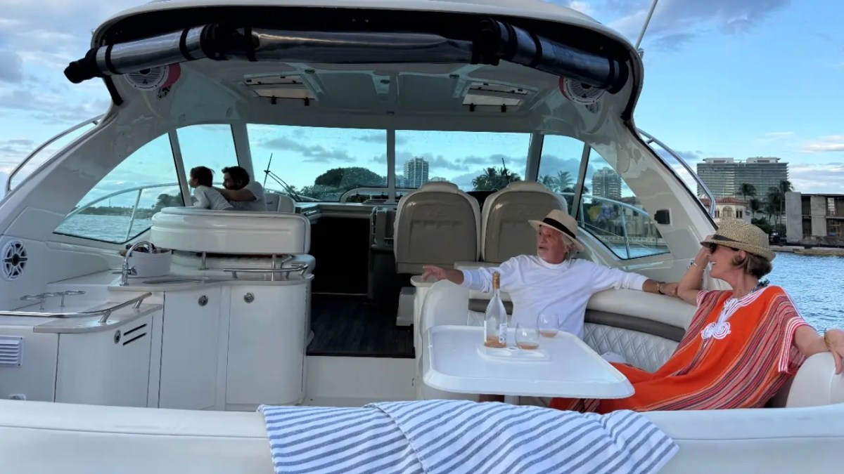 Two people relaxing on a yacht with drinks, two others sit at the helm, cityscape in the background.