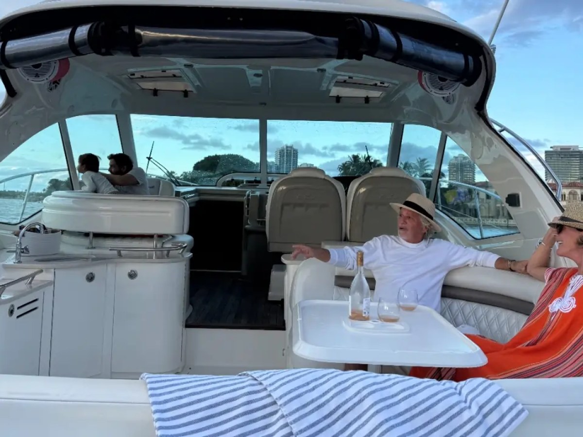 Two people relaxing on a yacht with drinks, two others sit at the helm, cityscape in the background.