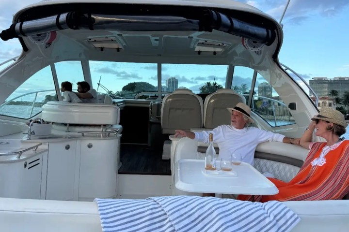 Two people relaxing on a yacht with drinks, two others sit at the helm, cityscape in the background.