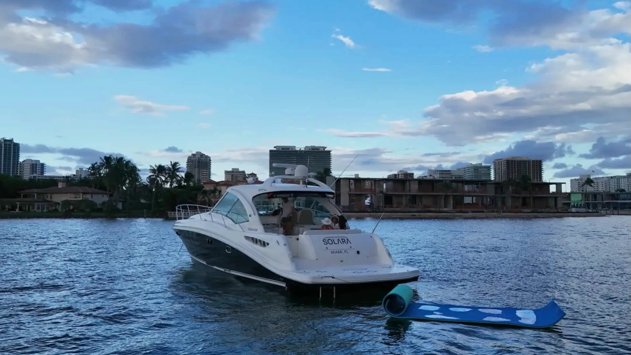 A boat named 'Solara' in a city harbor with a floating mat behind it, under a partly cloudy blue sky.