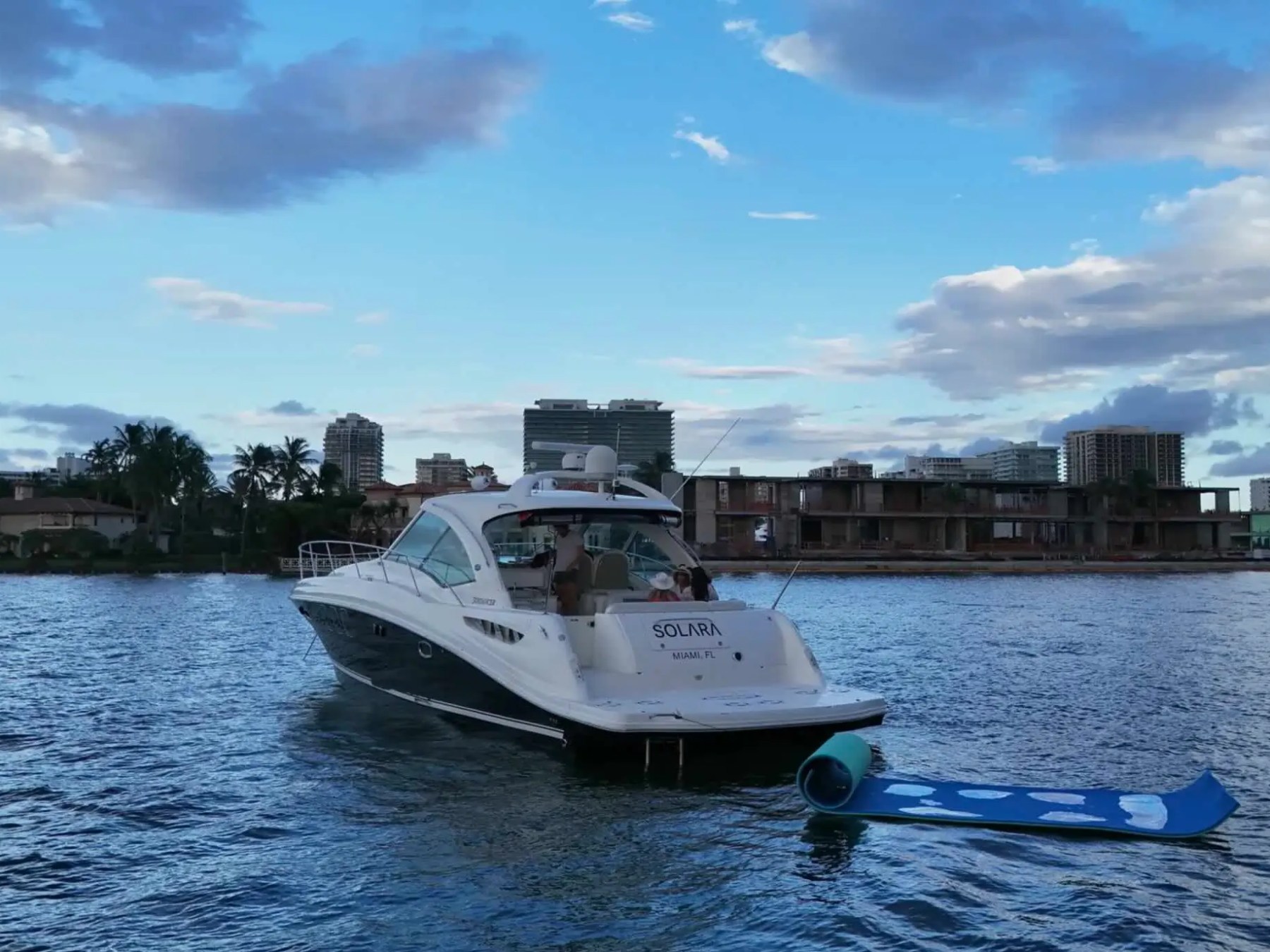 A boat named 'Solara' in a city harbor with a floating mat behind it, under a partly cloudy blue sky.