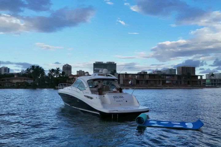 A boat named 'Solara' in a city harbor with a floating mat behind it, under a partly cloudy blue sky.