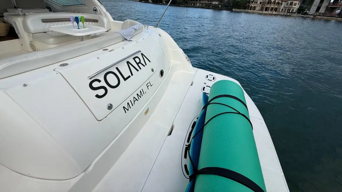 Back of a boat named 'Solara' with a rolled-up mat on deck, moored on calm water.