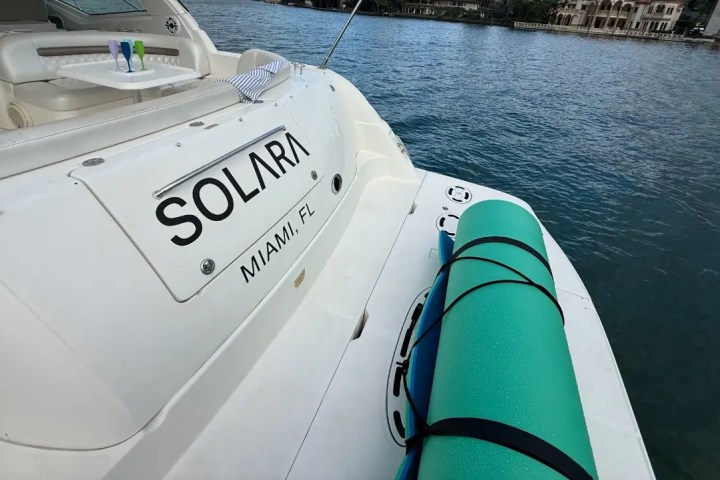 Back of a boat named 'Solara' with a rolled-up mat on deck, moored on calm water.