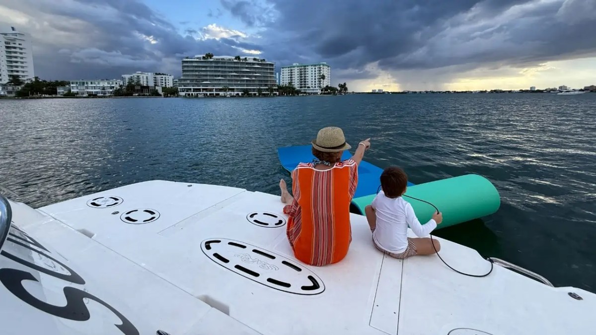 Two people sitting on a boat overlooking the water, with buildings and cloudy sky in the background.