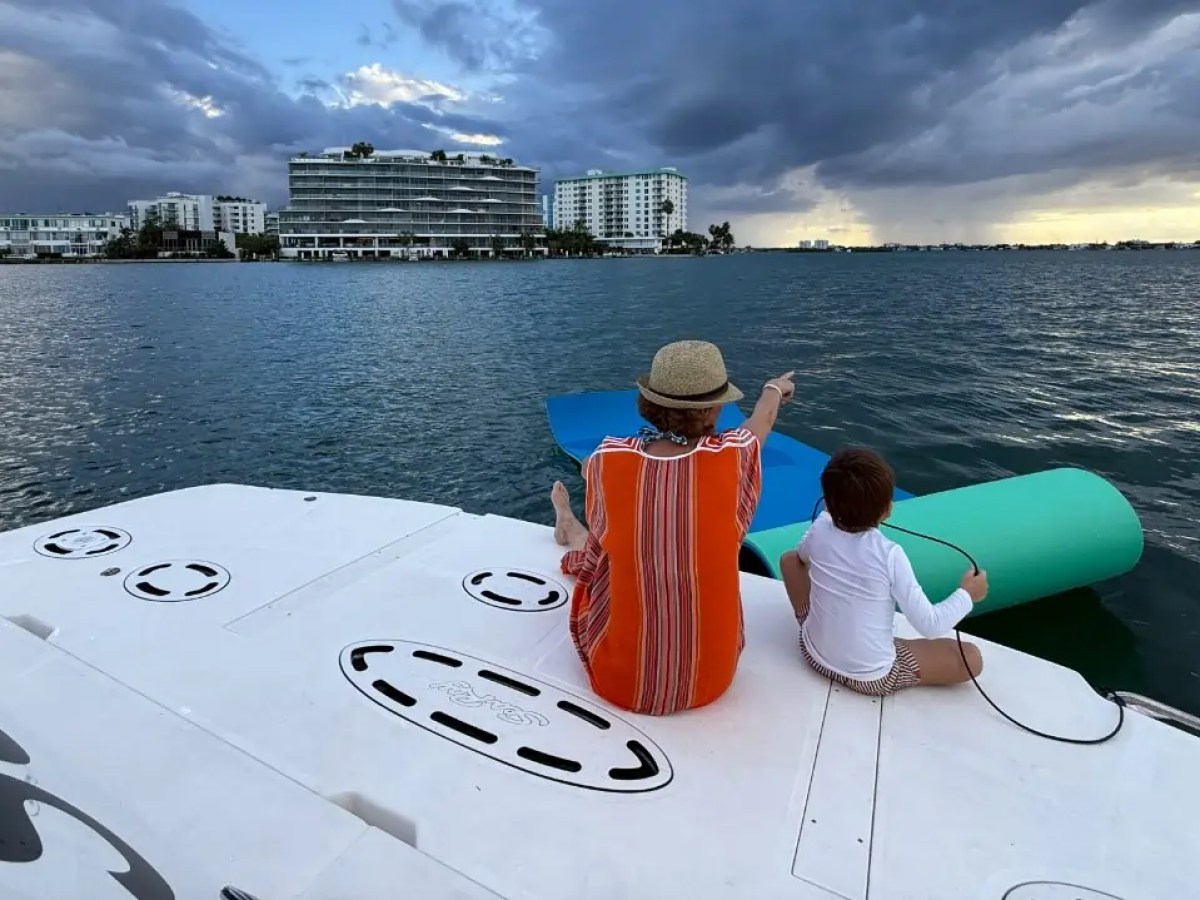 Two people sitting on a boat overlooking the water, with buildings and cloudy sky in the background.