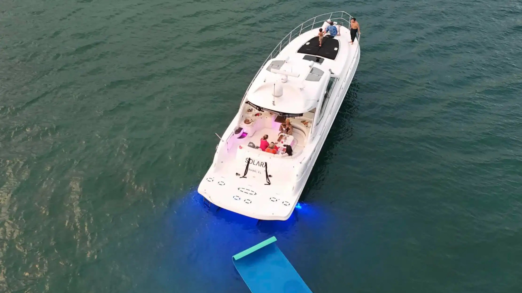 Overhead view of a white yacht with people on deck, blue water, and a floating mat trailing behind.