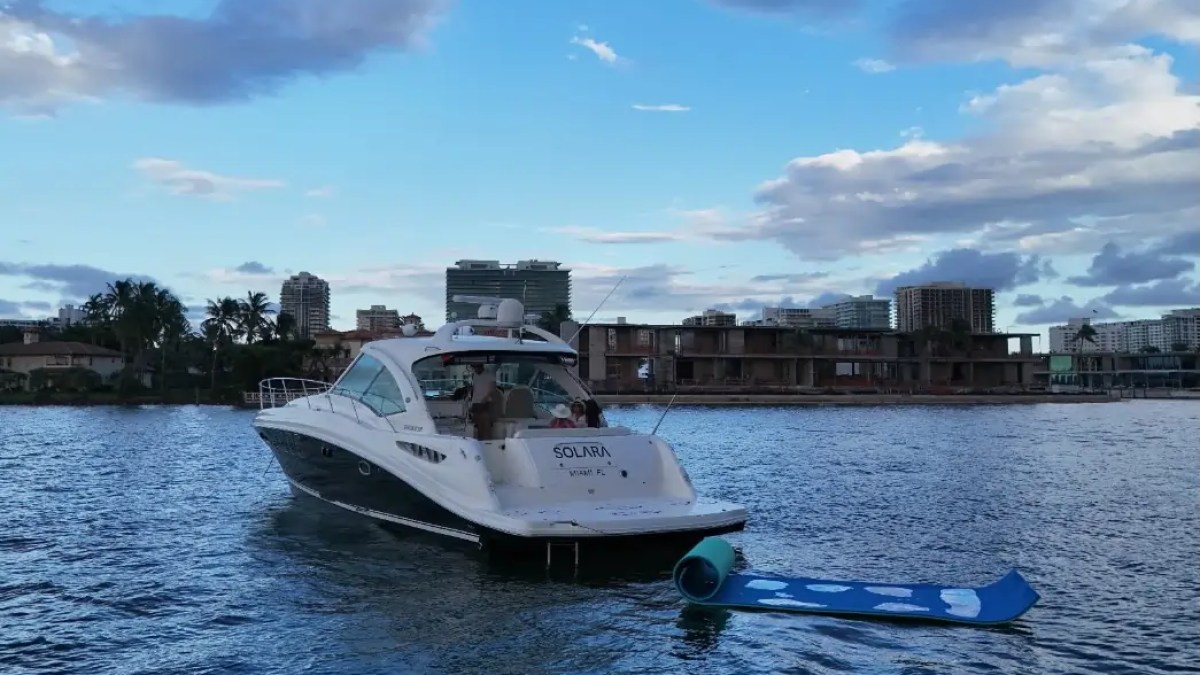 Yacht named Solara in water, skyline with buildings and palm trees in background, blue sky with clouds.