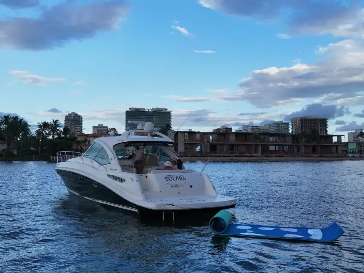Yacht named Solara in water, skyline with buildings and palm trees in background, blue sky with clouds.