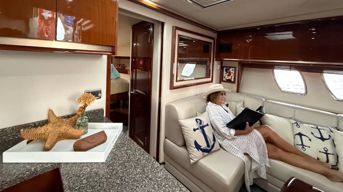 Woman in white dress and hat reads on a yacht sofa with nautical pillows, near a starfish decor.