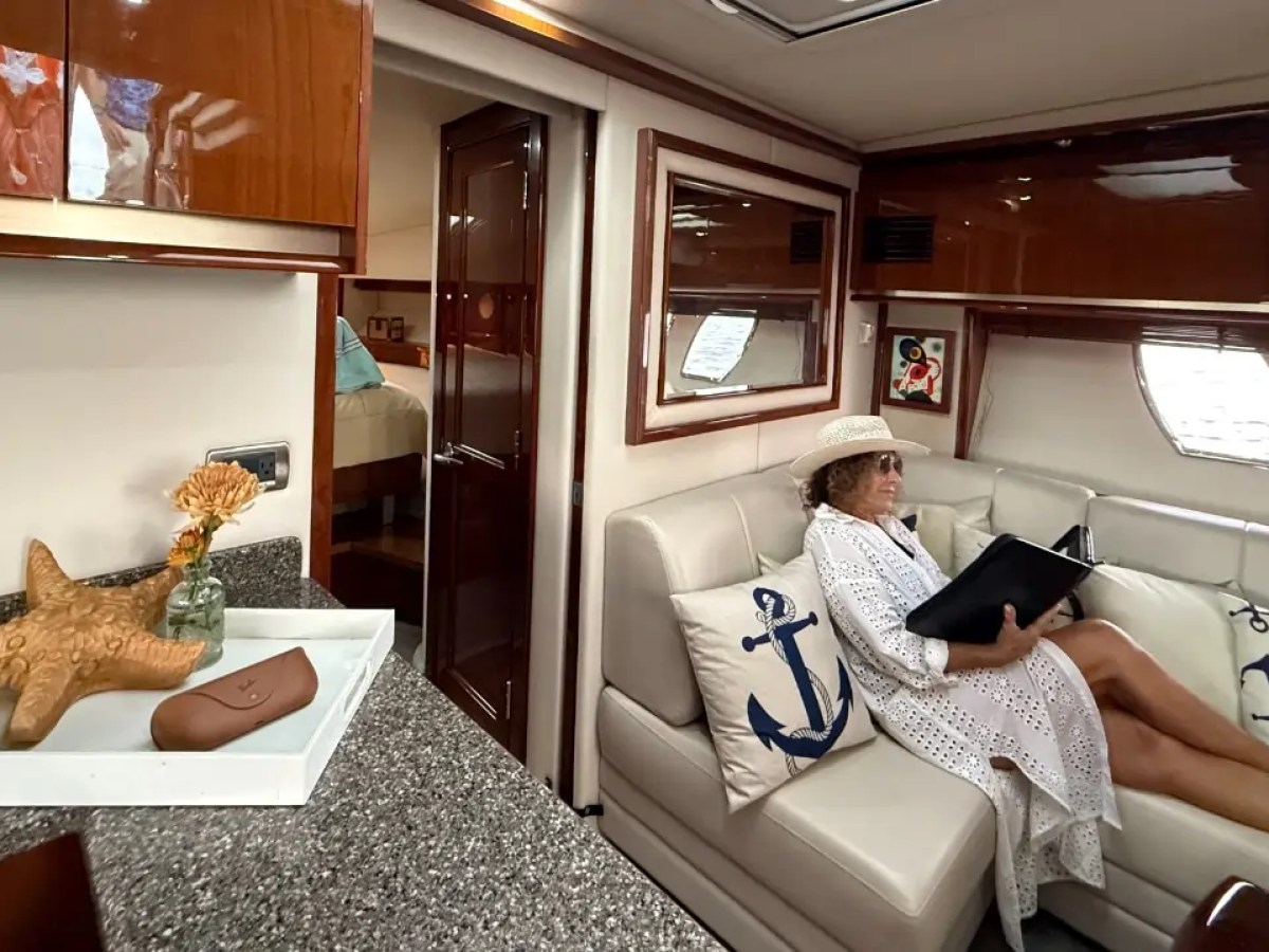 Woman in white dress and hat reads on a yacht sofa with nautical pillows, near a starfish decor.