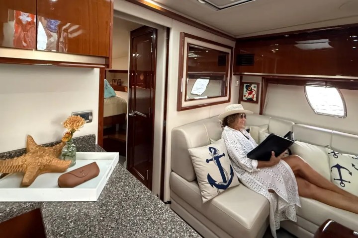 Woman in white dress and hat reads on a yacht sofa with nautical pillows, near a starfish decor.