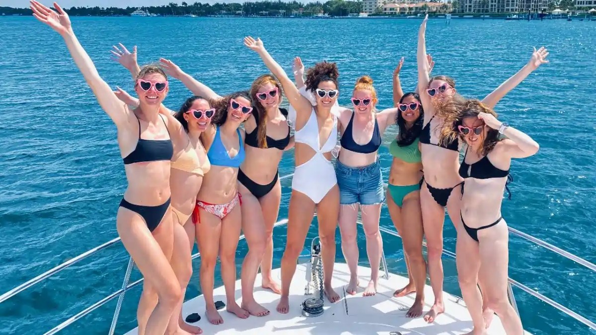 Group of women in swimsuits and heart-shaped sunglasses on a boat, arms raised.