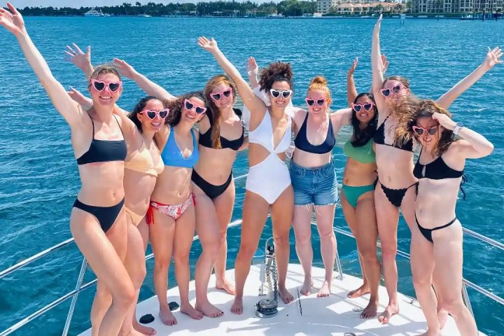 Group of women in swimsuits and heart-shaped sunglasses on a boat, arms raised.