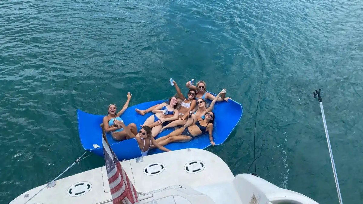 Group of people smiling on a blue float in the water near a boat with a flag.