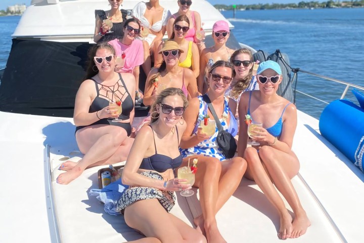Group of women relaxing on a yacht, wearing swimwear and sunglasses, holding drinks, with a scenic water view.