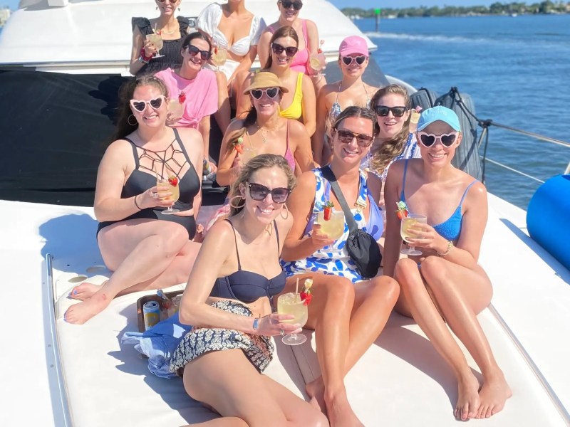 Group of women relaxing on a yacht, wearing swimwear and sunglasses, holding drinks, with a scenic water view.