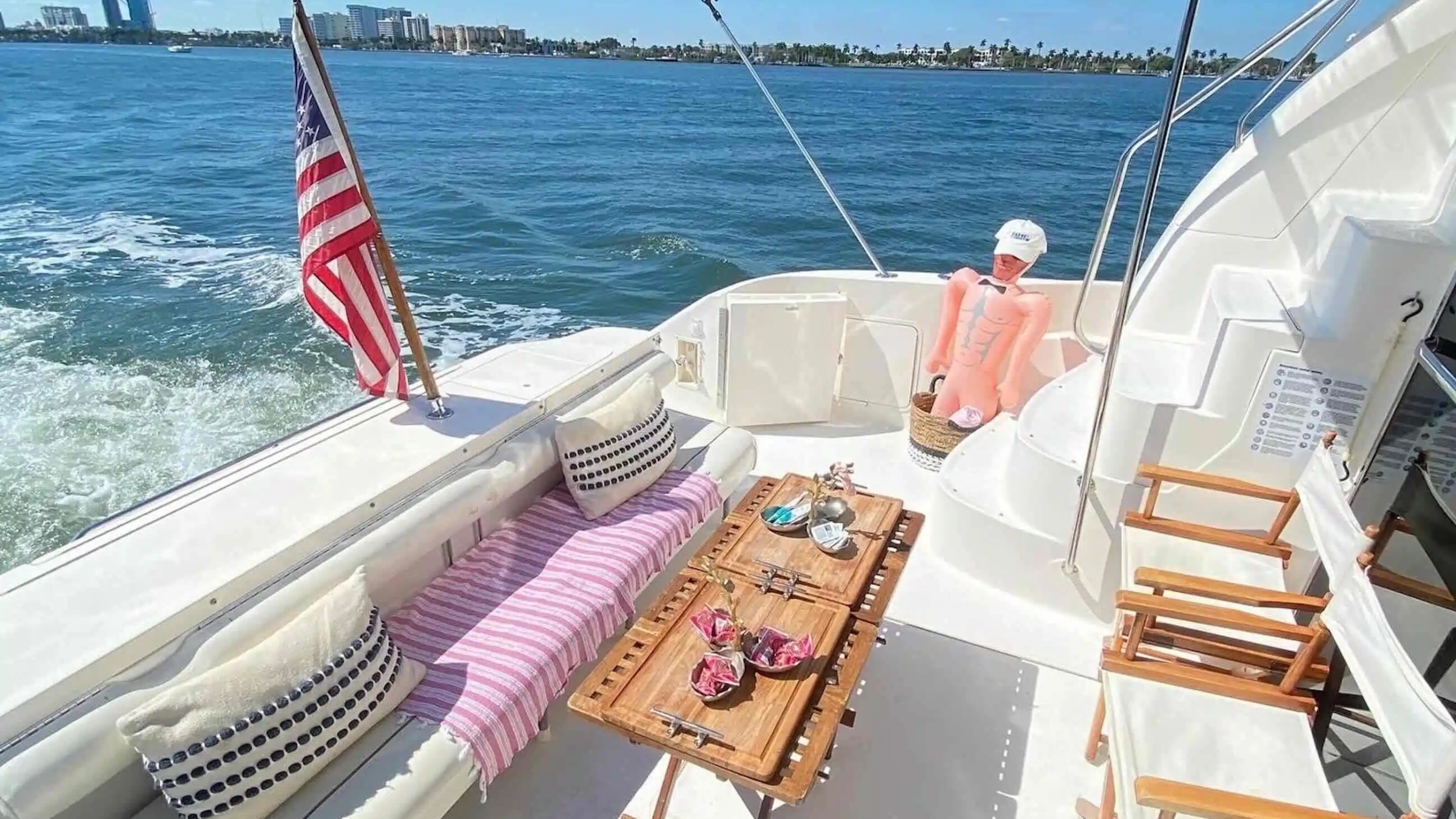 Boat deck with table, chairs, an inflatable figure, and an American flag, sailing on open water.