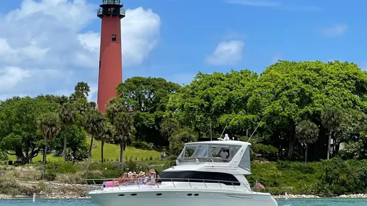 White yacht on water near red lighthouse and green trees under blue sky.