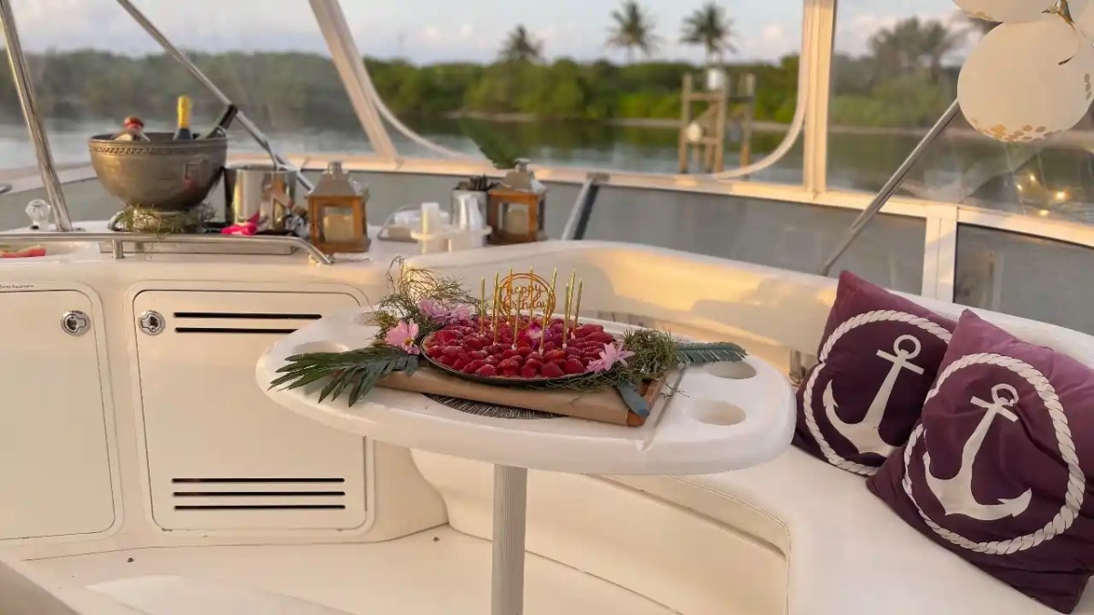 Yacht interior with fruit platter on table, anchor pillows, and background of water and trees.