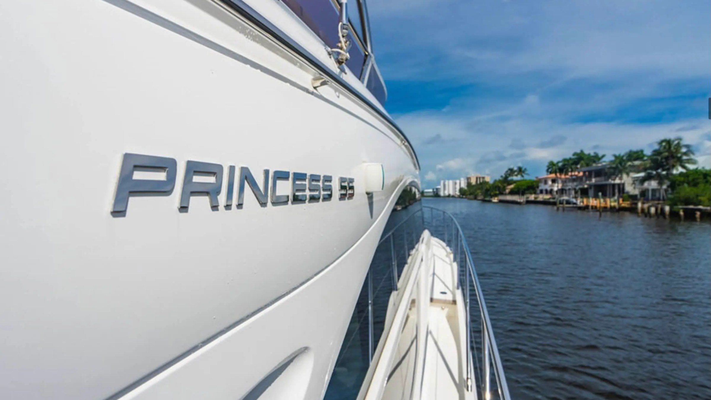 Side view of a white yacht with 'PRINCESS 55' text, cruising on a river lined with palm trees and houses.