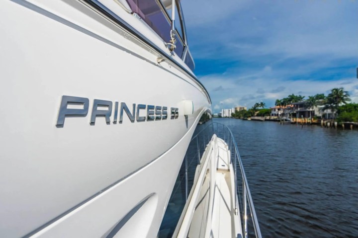 Side view of a white yacht with 'PRINCESS 55' text, cruising on a river lined with palm trees and houses.