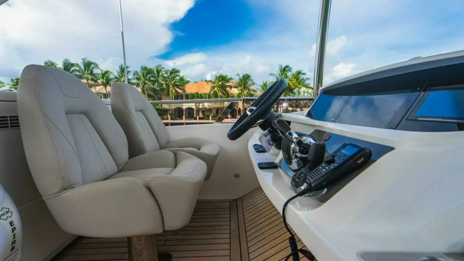 Luxury yacht interior with two beige seats and boat controls under a sunny sky with palm trees.