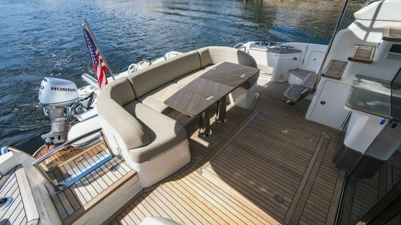 Boat deck with wooden flooring, curved seating, table, and an American flag near the water.