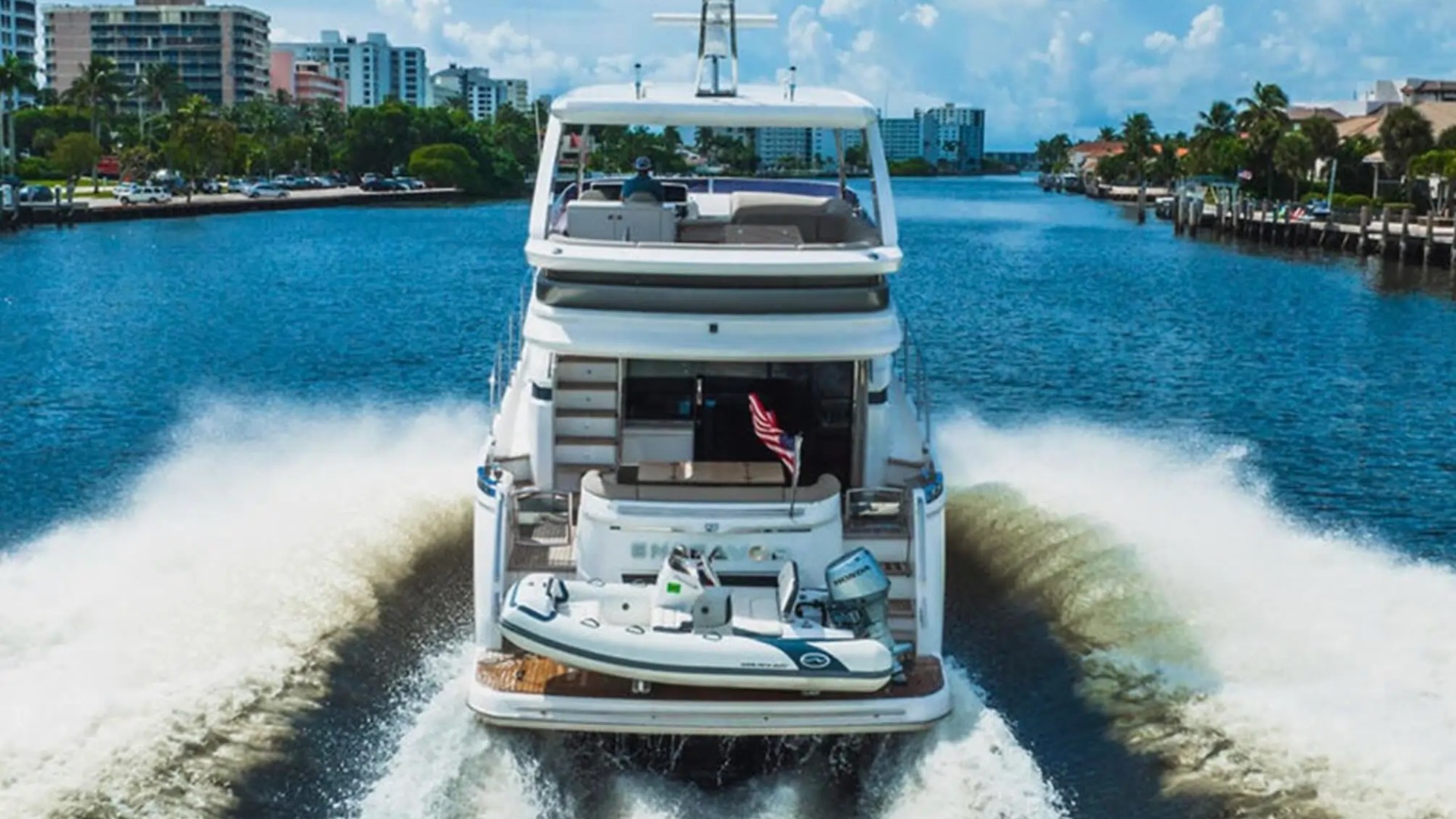 Large yacht cruising on a river with buildings and trees in the background.
