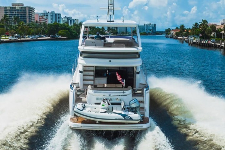 Large yacht cruising on a river with buildings and trees in the background.