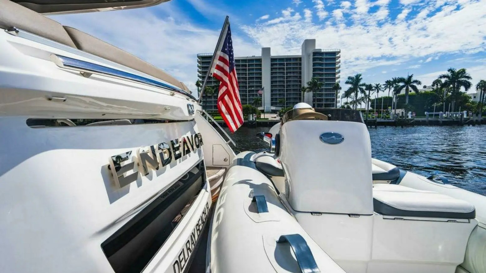 Luxury boat named Endeavor on water, U.S. flag, and building, palm trees in background.