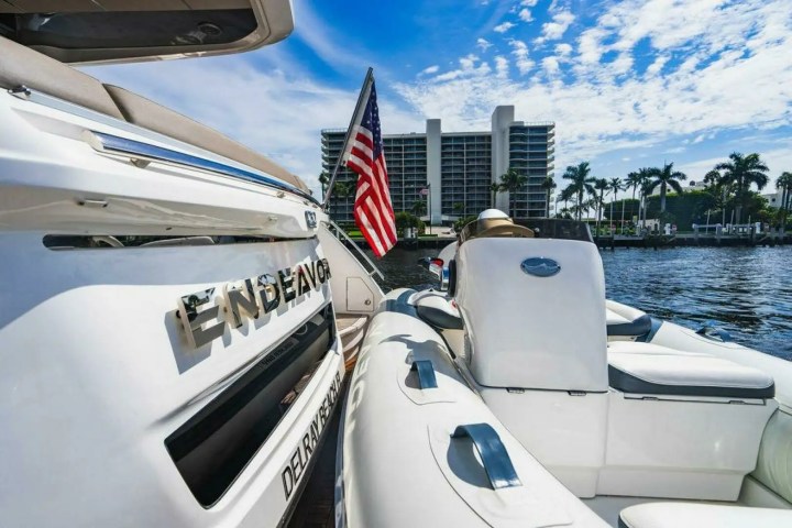 Luxury boat named Endeavor on water, U.S. flag, and building, palm trees in background.