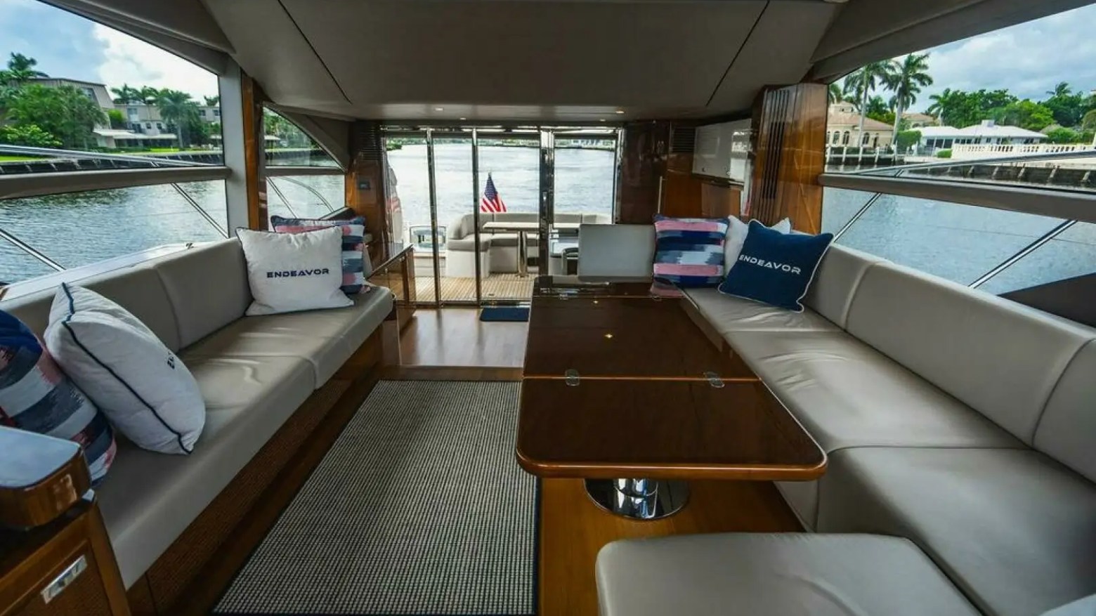 Yacht interior with beige seating, pillows, and a central wooden table, overlooking a waterfront view.