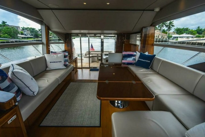 Yacht interior with beige seating, pillows, and a central wooden table, overlooking a waterfront view.