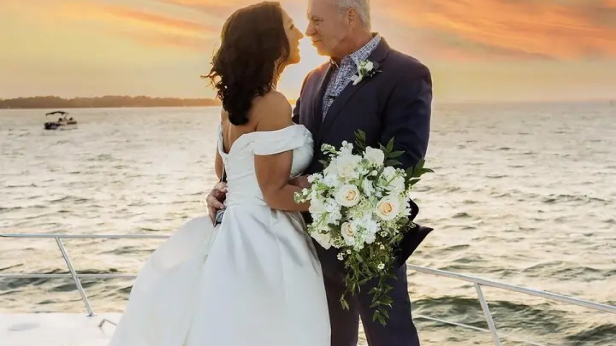 Couple in wedding attire embrace on a boat at sunset.