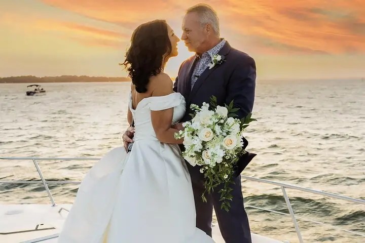 Couple in wedding attire embrace on a boat at sunset.