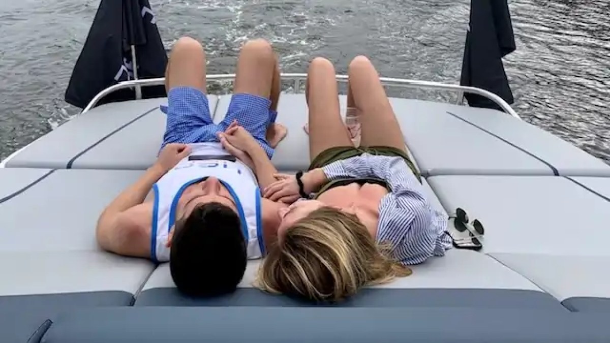 Two people relaxing on a boat deck, holding hands, with water visible behind them.