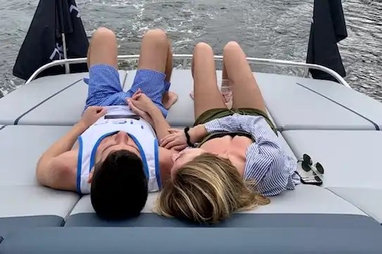 Two people relaxing on a boat deck, holding hands, with water visible behind them.