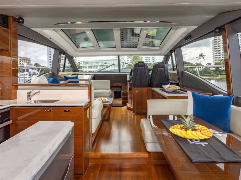 Luxurious yacht interior with wood floors, plush seating, and fruit platter on table. Large windows show a cityscape.