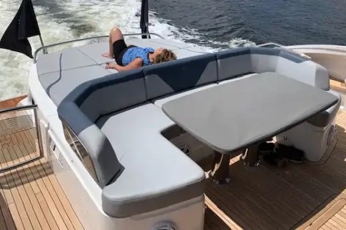 Person relaxing on yacht deck with cushioned seating and table, near water.